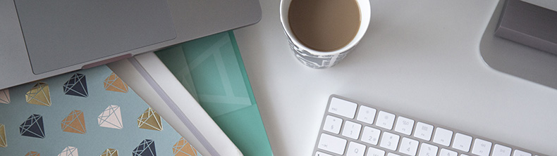 Työpöydällä on kahvikuppi, kannettava tietokone, näppäimistö ja muistiinpanovälineitä.